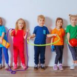 Auf diesem Bild sind 5 Kinder zu sehen, die dabei sind Sport zu machen. Sie haben eine Matte, einen Reifen und einen Ball dabei.