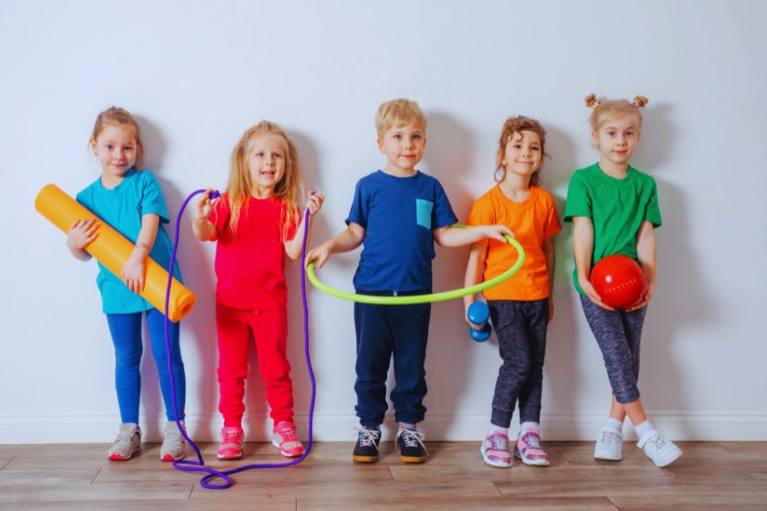 Auf diesem Bild sind 5 Kinder zu sehen, die dabei sind Sport zu machen. Sie haben eine Matte, einen Reifen und einen Ball dabei. 