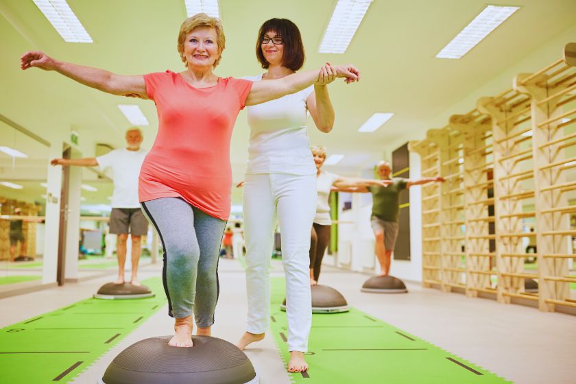 Eine ältere Dame macht den Einbeinstand auf einem Balance Trainer. Sie wird dabei von einer Trainerin unterstützt.  